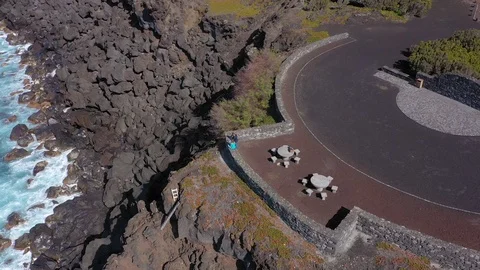 Two people look on ocean sitting at viewpoint above black stones. Varaduro do Stockbeeldmateriaal 117769346