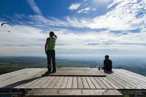 Two people looking at some flying paragliders, on a wood platform Stock Photos