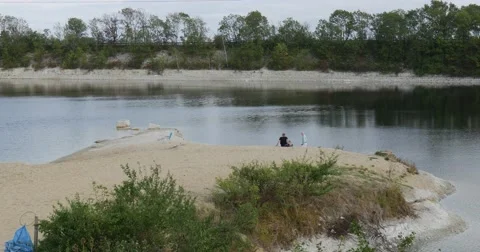 Two People Man is Sitting at River Lake Bank Sandy Bank of Peninsula Cloudy Stock Footage 54824579