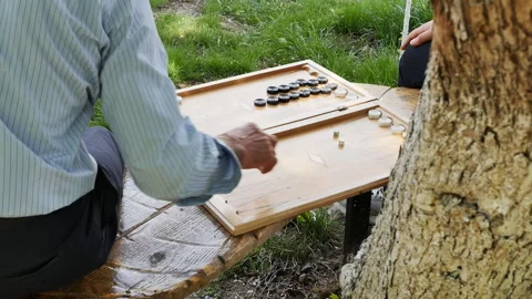 Two people play backgammon outdoors sitting on a bench. Senior man and young guy Stockbeeldmateriaal 194808067