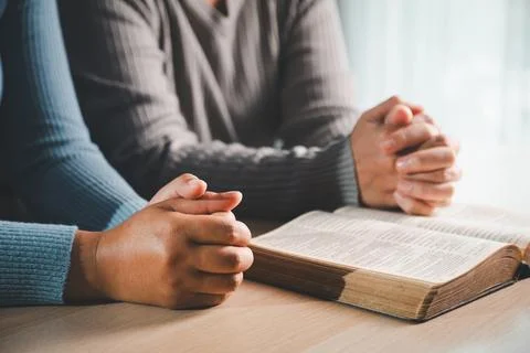Two people praying with folded hands beside open Bible on wooden table, sho.. Stock Photos
