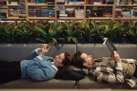Two people reading and using smartphone on couch in library Foto stock