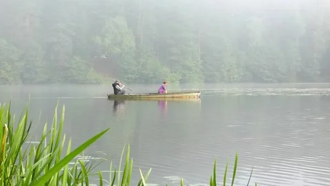 Two people row a boat on a quiet lake surrounded by trees in the morning mist wh Stock-Footage 325626467