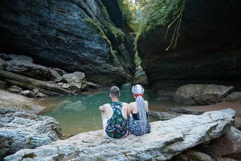 Two people sit on a large rock and look at the grotto, rocks and mountain river Stock Photos