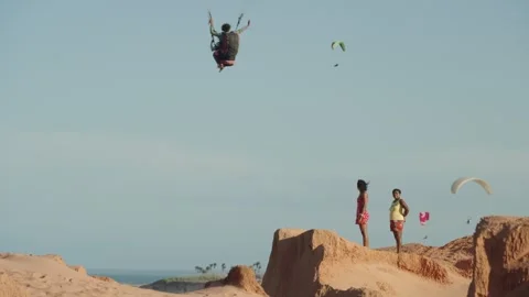 Two people stand on sandy coastal cliffs as colorful paragliders fly across a 動画素材 328587243