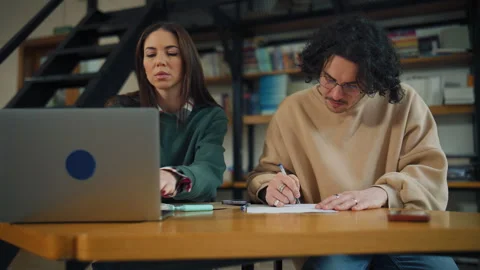 Two people study together in a library, observing a laptop while taking notes on Stock Footage 337325968