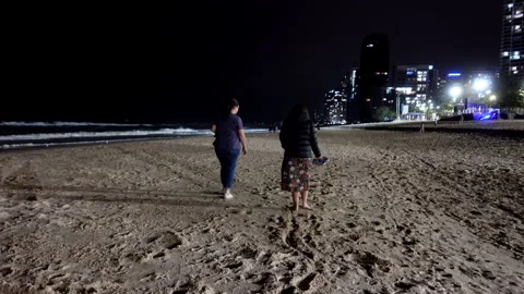 Two People Walking Barefoot on Beach at Night With City Lights Behind Stock Footage 329365910