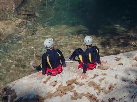 Two people in wetsuits and helmets sit on the rocky shore of a clear reservoir Stock Photos