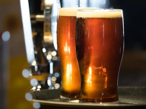 Two pints of dark coloured real ale in pint glasses with froth on bar top Stock Photos