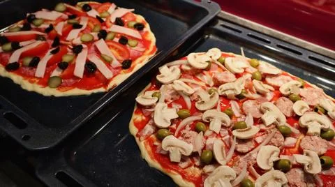 Two pizzas on baking trays, each topped with fresh ingredients like mushrooms. Stock Photos