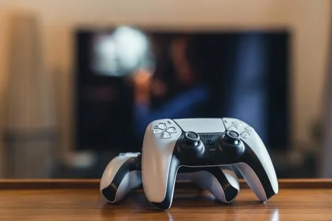 Two Playstation 5 (PS5) controllers at home, tv on background, selective focu Stock Photos