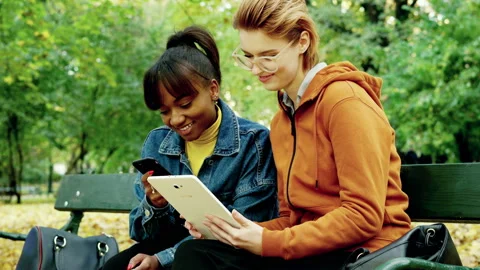 Two pretty girlfriends using smartphone and tablet in autumn park, 4K Stock Footage 144614337