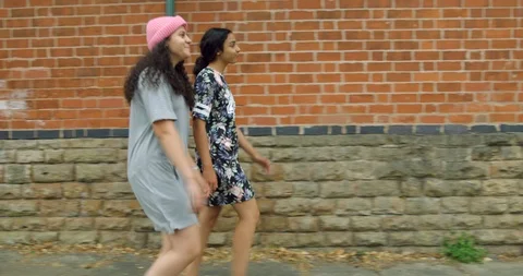 Two Pretty Mixed Race Young Girl Twins Walking Along a Street By Brick wall. Stock Footage 94550626