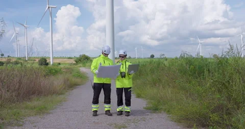 Two professional engineers examine map and laptop at wind turbine field discuss Stock Footage 325840142