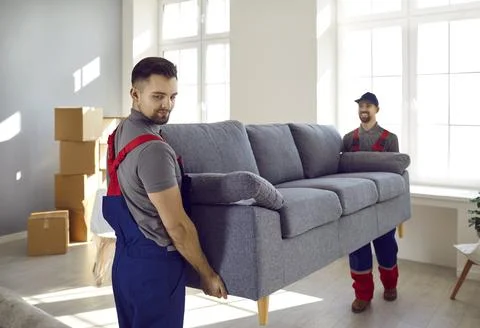 Two professional relocation service workers in overalls move sofa in customers Stock Photos