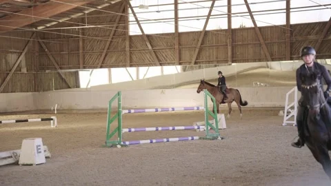 Two professional riders training horses for show jumping in a large, sandy Stock Footage 332650202