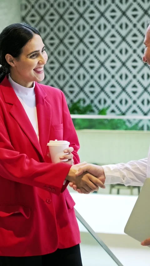 Two professionals shake hands in a modern office, one in a red blazer holdi.. Video stock 345518729