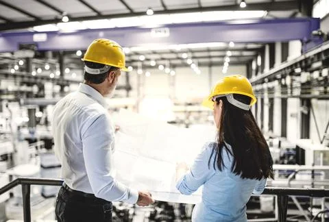 Two project managers standing in modern industrial factory, looking blueprints Stock Photos