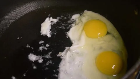 Two raw eggs, freshly cracked into a large black skillet cooking slowly. Video stock 339100968