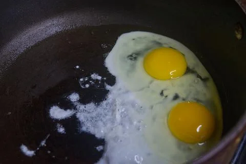 Two raw eggs, freshly cracked into a large black skillet cooking slowly. Stock Photos