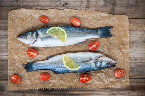 Two raw seabass fish with a lemon slice and cherry tomatoes on a rustic woo.. Stock Photos