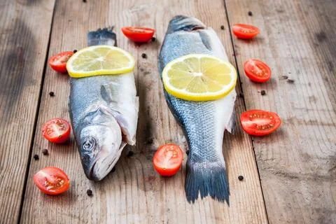 Two raw seabass fish with lemon and cherry tomatoes on a rustic wooden back.. Stock Photos