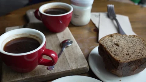 Two red coffee mugs with hot black coffee and a slice of banana bread are Stock Footage 336330523