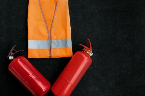 Two red fire extinguishers and an orange reflective road vest on a dark backg Stock Photos