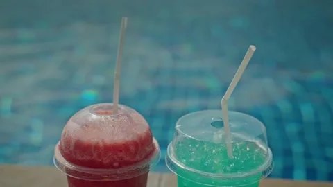 Two refreshing frozen drinks with straws, one red and one blue, sit by edge.. 스톡 동영상 334906091