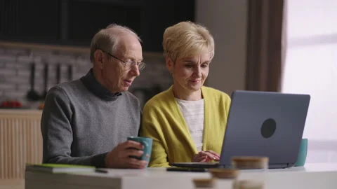 Two retirees are using laptop at home kitchen, surfing internet, shopping online Видео 153745242