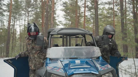 Two riders beside blue atv checking doors and windshield in snowy pine forest Stock Footage 332055946