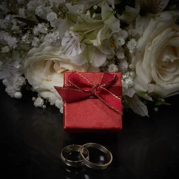 Two rings of different sizes, a red gift box lie on the background of a bouqu Stock Photos