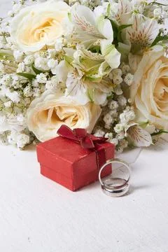 Two rings, a gift box in red color, a bouquet of white flowers on a white woo Stock Photos