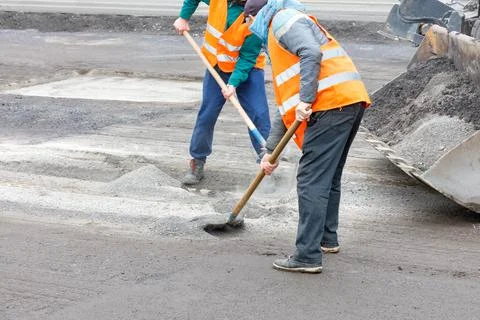 Two road workers rake old broken asphalt off the road and load it into a grad Stock Photos