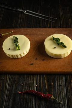 Two rounds of fresh cheese placed on a wooden cutting board with green herbs  Stock Photos