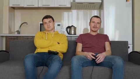 Two sad men throw their gamepads, rise from the couch and leave the frame Stock Footage 107652320