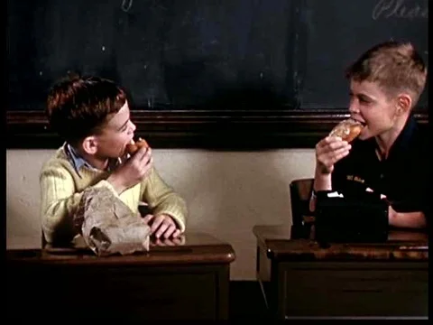 Two schoolboys eat donuts in their classroom and a presenter behind a desk, inƒ Stock Footage 75290889
