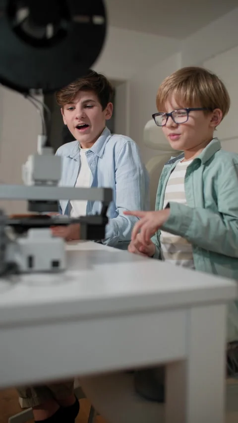 Two schoolboys at a white table chat, and watch a 3D printer in operation Stock Footage 335390443