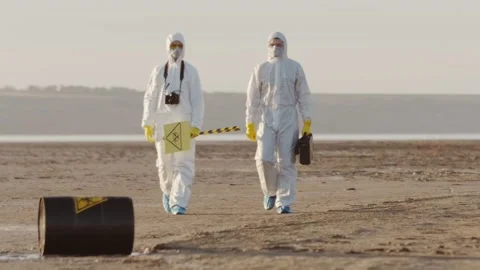 Two scientists in protective suits approach a black barrel. 動画素材 130660333