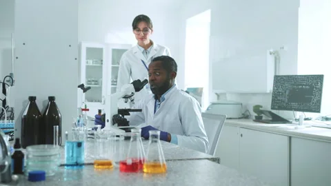 Two scientists in uniform looking under a microscope talking discuss wonder in Stock Footage 246750383