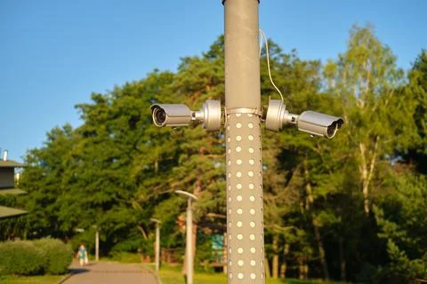 Two security cameras mounted on a metal pole in an outdoor park. Daytime sc.. Stock Photos