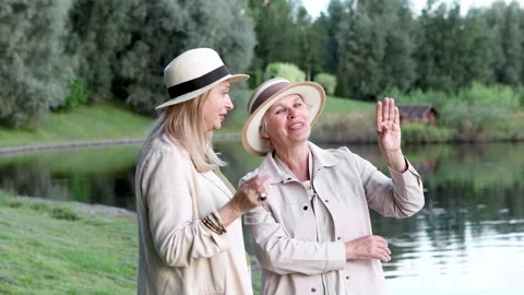 Two senior women talk while standing by lake in scenic landscape, enjoying soft Stock Footage 319016355