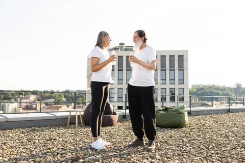 Two seniors converse while drinking water on an urban building rooftop Foto stock