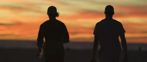 Two silhouetted surfers walking on the beach with a pet dog at sunset Stock Footage 111962706