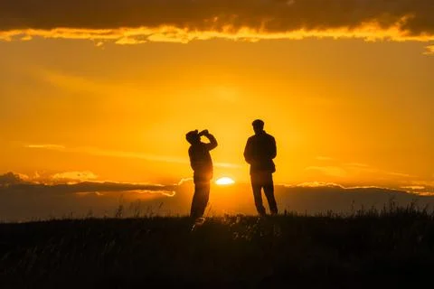 Two silhouettes of men talking and drinking a drink a jar at sunset or sunris 스톡 사진