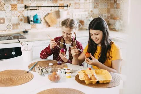 Two sisters are sitting at the table and painting Easter eggs in different Stock Photos