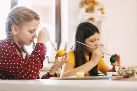 Two sisters are sitting at the table and painting Easter eggs in different Stock Photos