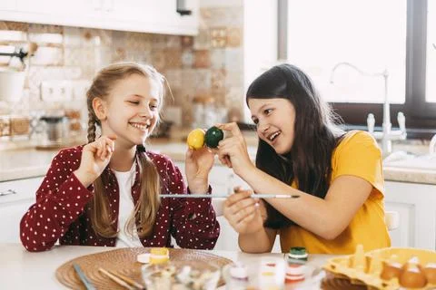 Two sisters are sitting at the table and painting Easter eggs in different Stock Photos