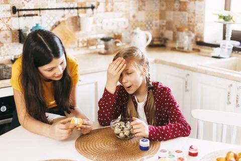 Two sisters are sitting at the table and painting Easter eggs in different Stock Photos