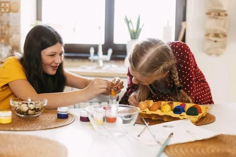 Two sisters are sitting at the table and painting Easter eggs in different Stock Photos
